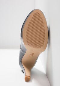 Chaussure à talon haut noire vue de la semelle, mettant en évidence la semelle beige texturée et le talon fin sur fond blanc.