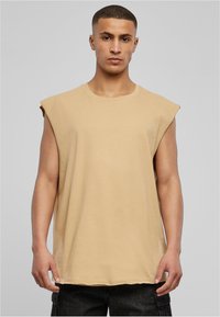 Beige mouwloze t-shirt van katoen. Ronde halslijn, losse pasvorm, schoudernaden lopen iets door, en de zoom eindigt ter hoogte van de heupen.