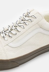 White suede sneaker with textured surface, white leather side stripe, black and white patterned laces, and a brown rubber sole.
