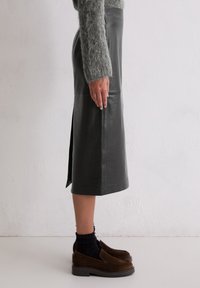 Ciemnozielona skórzana spódnica midi z bocznym rozcięciem, gładka faktura, długość do kolan i dopasowany krój, zestawiona z brązowymi zamszowymi butami wsuwanymi.