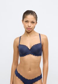 Σουτιέν σε ναυτικό μπλε με δαντέλα, με προσαρμοσμένα καλύμματα και ρυθμιζόμενους ιμάντες. Απαλό ύφασμα με κυματιστά άκρα, διακοσμημένο με ένα μικρό φιόγκο στο κέντρο.