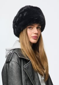 Chapéu de pele falsa preta com aba larga, apresentando uma textura suave e design arredondado. Usado com um hoodie cinzento e uma jaqueta de couro.