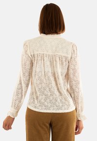 Blusa in pizzo color avorio con motivo floreale, maniche lunghe, yoke posteriore arricciato e polsini elasticizzati. Indossata con pantaloni in velluto a coste marroni.