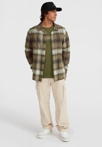Plaid flannel shirt in green and beige tones, layered over an olive t-shirt, paired with light beige cargo pants and white sneakers.