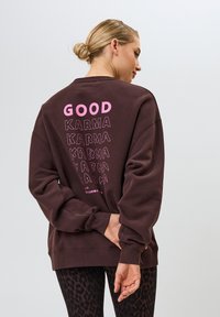 Sudadera marrón con cuello redondo. Presenta "GOOD" en texto rosa negrita, con la palabra "KARMA" en estilos variados superpuestos. Puños acanalados, corte relajado.