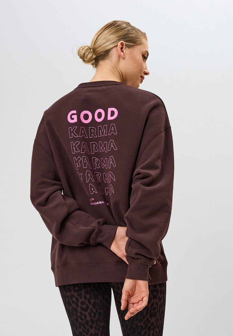 Sudadera marrón con cuello redondo. Presenta "GOOD" en texto rosa negrita, con la palabra "KARMA" en estilos variados superpuestos. Puños acanalados, corte relajado.