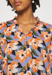 Blouse à motif floral bleu clair, ornée de grandes fleurs orange et blanches. Col à volants, fermeture à boutons, tissu texturé. Boutons nacrés.