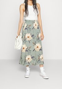 Falda maxi floral verde con flores crema y marino, cinturilla elástica y diseño fluido. Combinada con una camiseta de tirantes blanca y zapatillas.