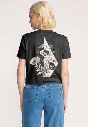 Black cropped t-shirt featuring a floral design in beige on the back, with a geometric accent. Paired with light blue high-waisted jeans.