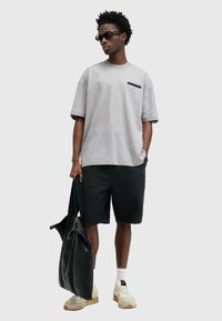 Mann som har på seg solbriller, grå oversized t-skjorte, svarte shortser, hvite joggesko, og bærer en stor svart lærveske mot en ensfarget bakgrunn.