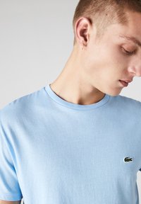 Camiseta de algodón azul claro con cuello redondo y un pequeño logo de cocodrilo bordado en verde en el lado izquierdo del pecho. Textura suave.