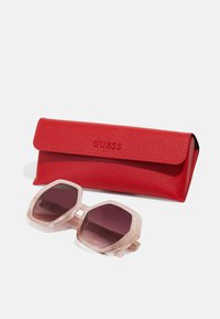 Red leather sunglasses case with embossed "GUESS" logo, paired with hexagonal sunglasses featuring translucent light pink frames and dark tinted lenses.