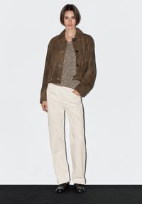 Hnědá semišová zkrácená bunda s černými knoflíky, béžový svetr s texturou, krémové kalhoty s širokými nohavicemi a černé boty na jednobarevném pozadí.