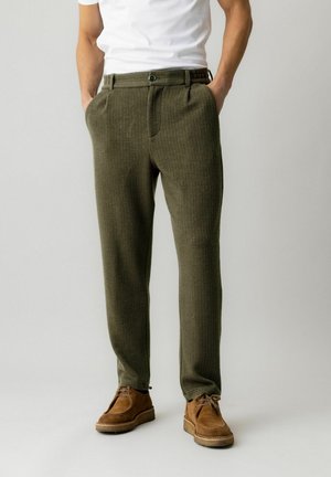 Hombre con pantalones texturizados de color verde oliva, zapatos de ante marrones y una camisa blanca de manga corta, de pie con las manos en los bolsillos.