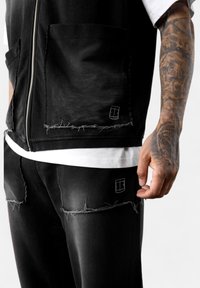 Schwarzes, ausgewaschenes Stoffoutfit mit einer Reißverschlussweste mit großen Taschen und passenden Hosen mit fransigen Kanten und dezenten Logo-Akzenten.