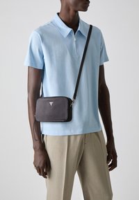 Zwarte schoudertas met een ritssluiting, rechthoekige vorm, textuur materiaal en een zilveren logo accent, gedragen over een lichtblauwe polo.