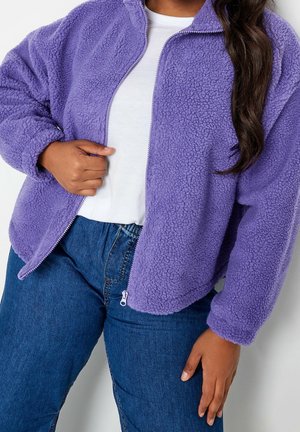 Person trägt eine hellviolette, flauschige Jacke mit Reißverschluss über einem weißen Shirt und blauen Jeans, wobei eine Hand den Reißverschluss der Jacke hält.