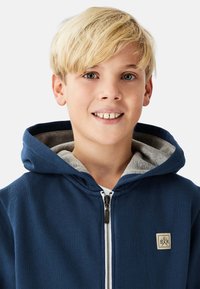 Marineblå lynlåshættejakke med gråt indre for, med et lille logo på brystet og en kængurulomme.