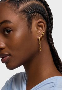 Los pendientes de aro dorados presentan diferentes formas, incluyendo un lazo, un óvalo y un diseño de candado, exhibiendo una textura metálica suave.