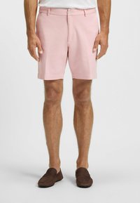 Light pink shorts made of cotton-stretch fabric with side pockets, a button closure, and a straight fit. Brown loafers on feet.