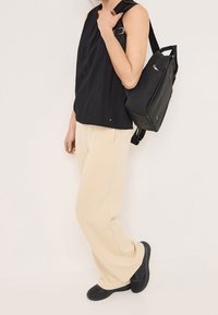 Kvinna iklädd svart ärmlös topp, beige vida byxor, svarta sneakers och bärande en svart ryggsäck på ena axeln mot en enfärgad bakgrund.