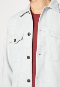 Lys grå knappet uldjakke med to forlommer, båret over en maroon crew-neck T-shirt på en person.