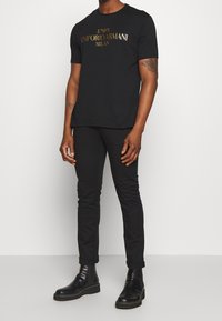 Svart kortärmad skjorta med guld "Emporio Armani"-logotyp, tillsammans med svarta slim fit-byxor och svarta ankelboots, i en enkel, minimalistisk design.
