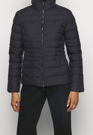 Black quilted zip-up jacket with high collar worn over black pants, showing upper body and arms against light background.