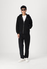 Hanorac negru cu fermoar, cu buzunare frontale, purtat peste un tricou alb. Asortat cu pantaloni negri și adidași albi. Modelul poartă ochelari de soare.