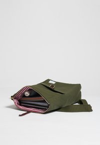 Bolsa de lona verde oliva con apertura con cremallera y un broche dorado, forrada con tela de rayas rojas y blancas, que contiene cuadernos y un tubo.