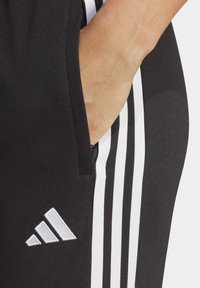 Schwarze Sporthose mit weißen Streifen an den Seiten und gesticktem Adidas-Logo. Der Stoff hat eine glatte Textur.