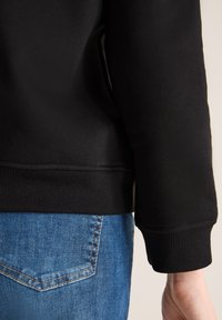 Sudadera negra con puños y dobladillo acanalados, textura suave, combinada con jeans de mezclilla azul. Muestra detalles de la manga y la cinturilla.