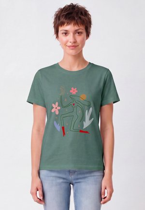 Femme aux cheveux courts portant un t-shirt vert avec une figure abstraite, des fleurs, des étoiles et des formes en rouge, rose, violet et blanc.