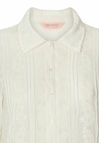 Chemise blanche à boutons, fabriquée en tissu léger, présentant un motif texturé et des rayures verticales. Comprend un col et des boutons ressemblant à des perles.