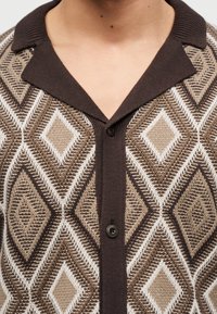 Καφέ και μπεζ πλεκτή ζακέτα με διαμάντι μοτίβο, με σκούρο καφέ γιακά και κουμπιά, φορεμένη από άτομο με εμφανή λαιμό και άνω μέρος στήθους.