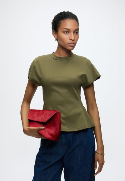 Γυναίκα με κοντά σγουρά μαλλιά που φοράει μπλούζα με κοντά μανίκια σε ελαιοπράσινο χρώμα και σκούρο παντελόνι, κρατώντας μια κόκκινη υφασμάτινη τσάντα χειρός.
