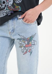 Jeans in denim azzurro chiaro con un dettagliato design di strass che presenta uccelli, cuori e spirali sulla coscia sinistra. Tasca anteriore visibile.