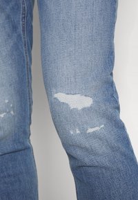 Světle modré džíny z denimu mají opotřebované detaily s třepícími se záplatami nad a pod kolenem, zobrazující texturovanou tkanou látku.