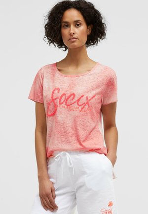 Woman with curly hair wearing a pink short-sleeve shirt with text and white drawstring shorts, standing with one hand in pocket.