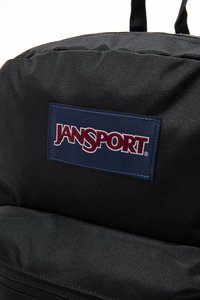Sac à dos en tissu noir avec un logo JanSport bien visible sur une étiquette rouge et bleue, avec une surface texturée et des détails de couture.