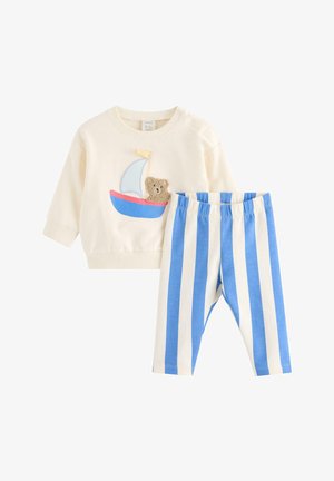 Baby-Shirt mit langen Ärmeln in Cremefarben, mit einem Segelboot- und Teddybär-Aufnäher, kombiniert mit blau-weiß gestreiften Hosen mit vertikalen Streifen.
