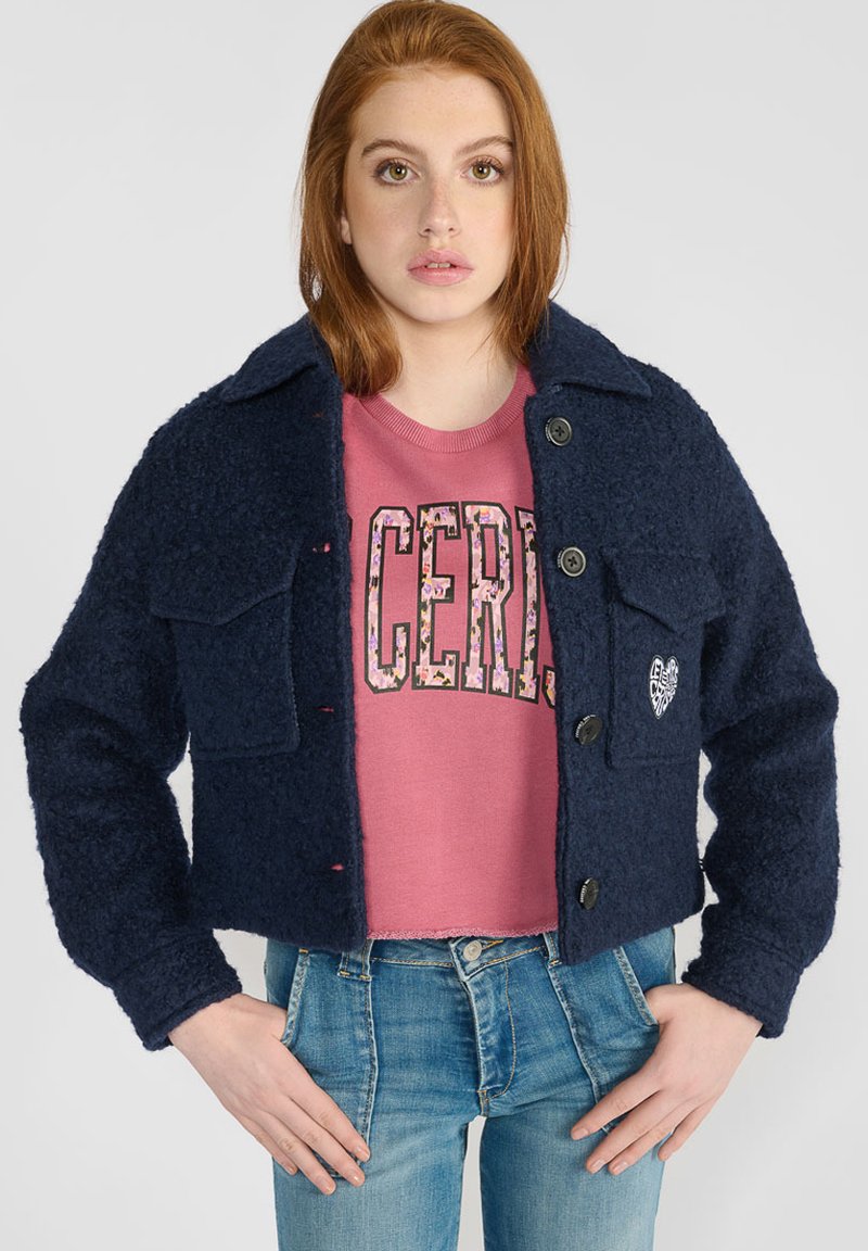 Veste cropped bleu marine en tissu texturé, dotée de deux poches poitrine, d'une fermeture à boutons et d'un détail brodé en forme de cœur.