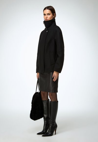 Sort oversized frakke med høj krave, kombineret med et tætsiddende sort lædernederdel og knælange sorte stiletstøvler. Fremhævet med en fluffy sort taske.