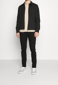 Giacca a strisce nere con zip, camicia beige sotto, pantaloni neri e sneakers grigie con lacci bianchi. Sfondo semplice.