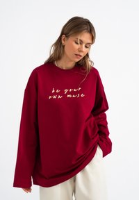 Bordeaux oversized sweatshirt met gouden tekst "wees je eigen muze." Heeft een ronde hals, verlaagde schouders en een zachte, gladde textuur.