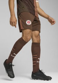 Shorts de football marron avec un patch logo, associés à des chaussettes hautes marron à rayures. Baskets de football noires avec crampons.