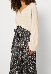 Haut en tricot côtelé beige avec un décolleté en V et des détails de boutons, accompagné d'une jupe noire avec un motif d'étoiles blanches et dorées. Manches longues, texture douce.