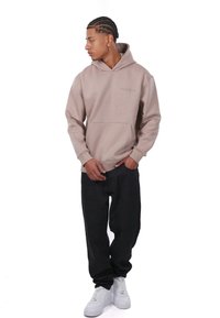 Tan hoodie med en frontlomme og subtilt logo, parret med løse sorte bukser og hvide sneakers, som præsenterer en afslappet pasform.