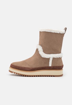 Barna velúrbőr csizma fehér prémmel, többszintű kialakítással és texturált gumitalpával. Hátsó húzófüllel a könnyű felhúzás érdekében.