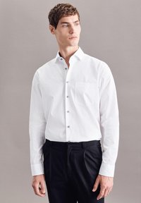 Witte blouse met lange mouwen, een button-down kraag, een borstzak en zilveren knopen. Draag het met zwarte broek tegen een neutrale achtergrond.